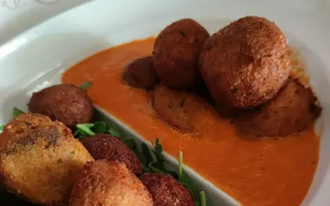 Polpette di pane dorate e croccanti, servite su una salsa rossa cremosa e guarnite con rucola fresca.