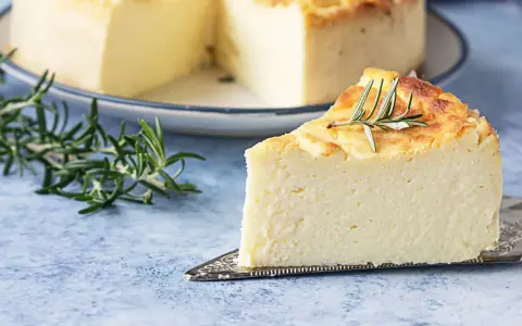 Una fetta di torta di ricotta soffice e dorata, guarnita con rosmarino, su un piatto da torta argentato.