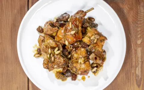 Pezzi di coniglio alla ligure, cotti con olive, pinoli e erbe aromatiche, serviti su un piatto bianco.