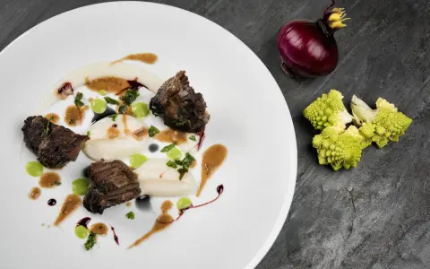 Succulenta guancia di manzo tenerissima, servita su purè bianco con gocce verdi e salsa bruna. Accanto, cipolla rossa e cavolfiore romanesco.