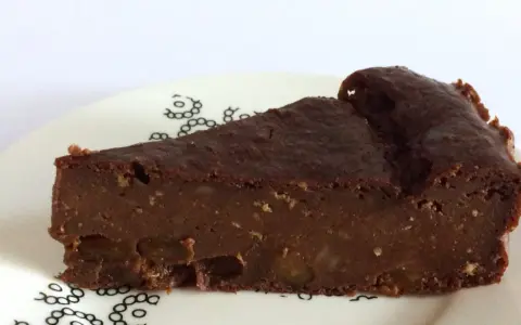 Una fetta di torta di pane al cioccolato, umida e ricca, con pezzi di frutta secca. La torta di pane ricetta della nonna, servita su un piatto bianco con decorazioni nere.