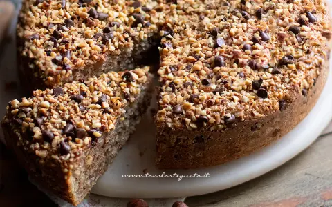 Una deliziosa torta di nocciole, ricoperta di granella di nocciole e gocce di cioccolato, con una fetta tagliata.