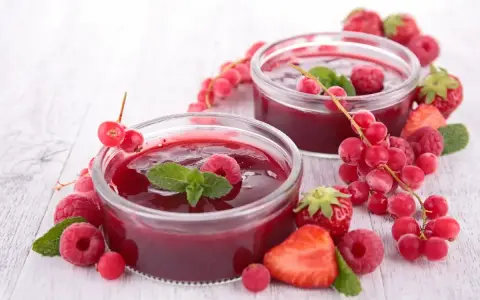 Due coppette di coulis di lamponi, guarnite con lamponi freschi, ribes e foglie di menta, circondate da fragole e frutti di bosco.