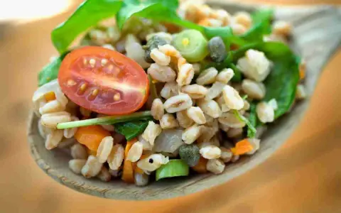 Un cucchiaio di farro condito con pomodorini, capperi e verdure fresche. Un'idea gustosa su come condire il farro per un pasto leggero e nutriente.