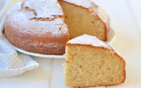 Una fetta di torta di banane soffice, cosparsa di zucchero a velo, accanto alla torta intera su un piatto bianco.