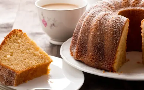 Una fetta di torta al latte soffice e dorata, cosparsa di zucchero a velo, accanto a una ciambella intera e una tazza di tè.