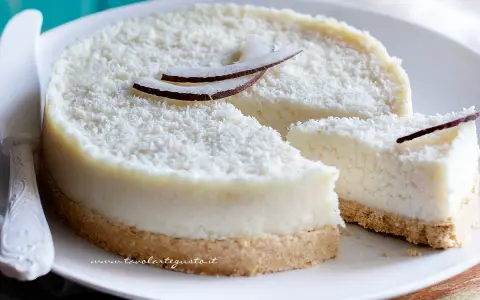 Delizioso cheesecake al cocco, ricoperto di cocco grattugiato e decorato con scaglie di cocco fresco. Una fetta è già stata tagliata.