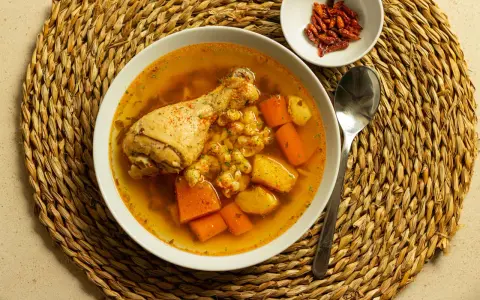 Un confortante brodo di pollo con pezzi di pollo, carote e patate, servito su una tovaglietta di paglia.
