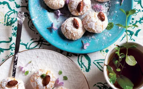 Biscotti al cocco spolverati di zucchero a velo, guarniti con mandorle e fiori viola, accanto a una tazza di tè alla menta.