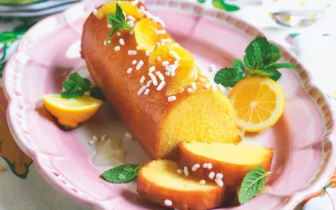 Deliziosa torta al limone glassata, decorata con fette d'arancia, zuccherini e menta. Servita su un piatto rosa con spicchi di limone.