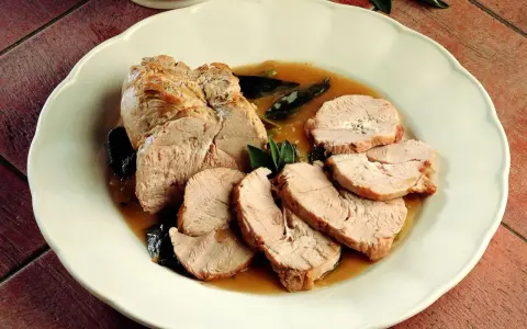 Delizioso arrosto di tacchino affettato, servito con salsa e foglie di salvia su un piatto bianco.