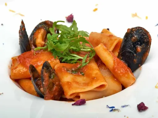 Paccheri allo scoglio con cozze e pomodoro, guarniti con rucola fresca e petali di fiori.