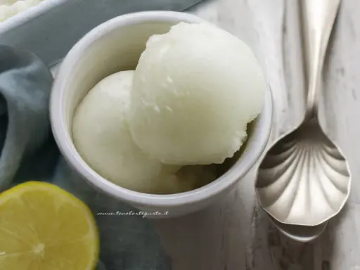 Coppetta con due palline di sorbetto al limone senza albume, accanto una fetta di limone e un cucchiaio decorato.