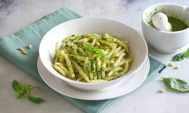 Un piatto di pasta con pesto fresco, guarnito con basilico e pinoli. Accanto, un mortaio con altro pesto.