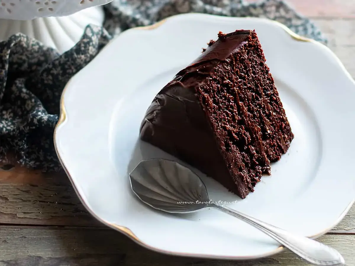 Una fetta di torta al cioccolato, umida e ricca, su un piatto bianco con bordo dorato. Accanto, un cucchiaino d'argento.