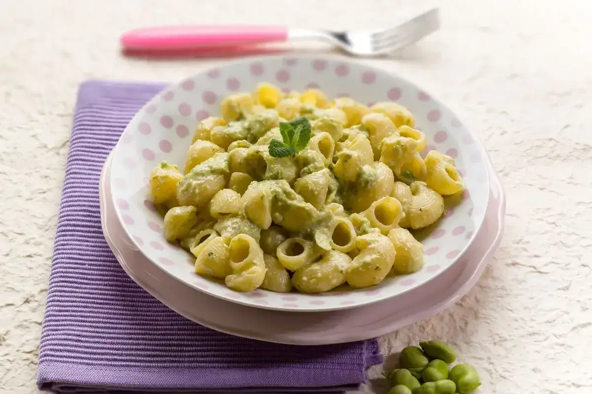 Piatto di pasta con le fave, condita con una crema verde e guarnita con menta. Un contorno di fave fresche completa la scena.