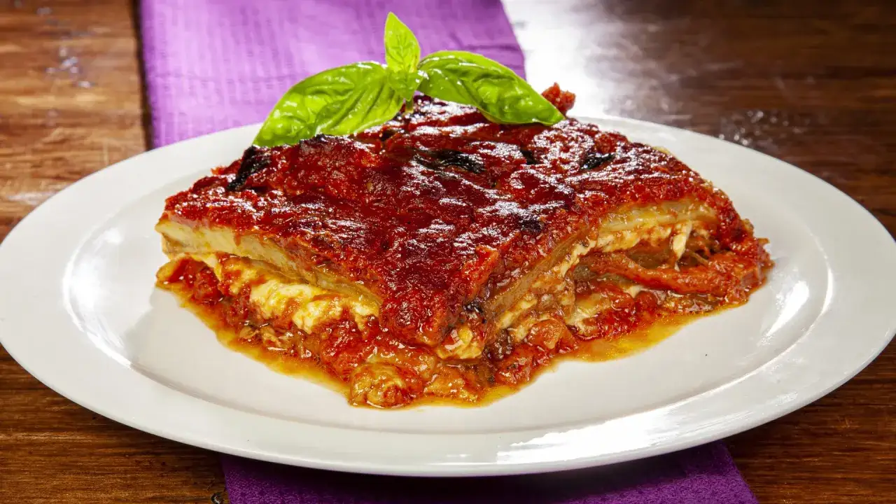 Una fetta di parmigiana di melanzane, ricca e succulenta, guarnita con foglie di basilico fresco, su un piatto bianco.