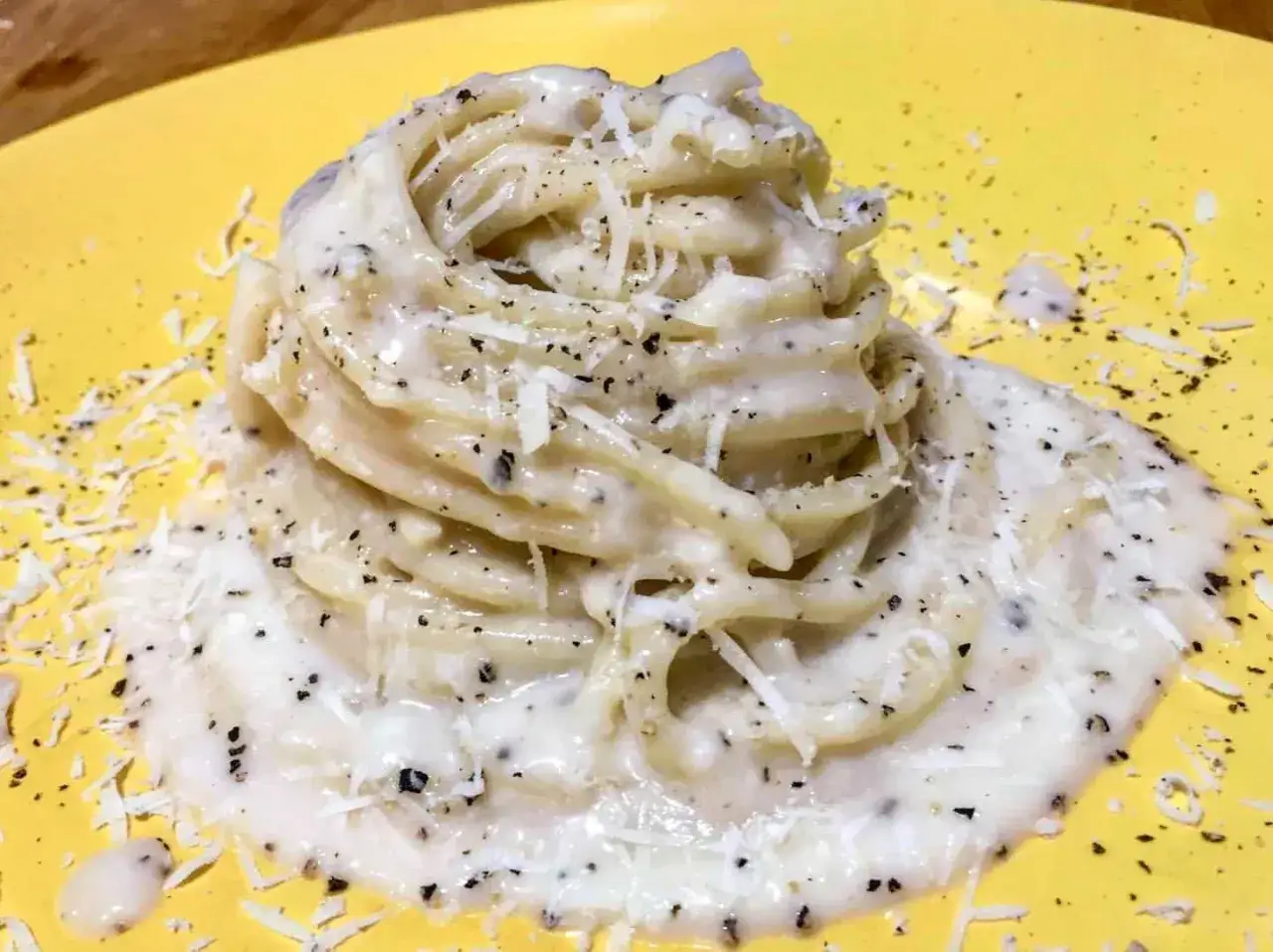 Un piatto di spaghetti cacio e pepe cremosi, cosparsi di pecorino grattugiato e pepe nero macinato fresco, serviti su un piatto giallo brillante.