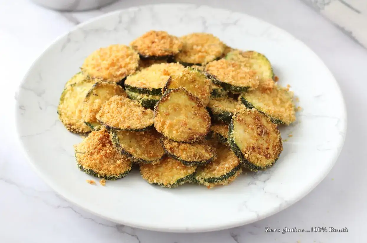 Fettine di zucchine impanate e cotte nella friggitrice ad aria, dorate e croccanti, servite su un piatto bianco con venature grigie.