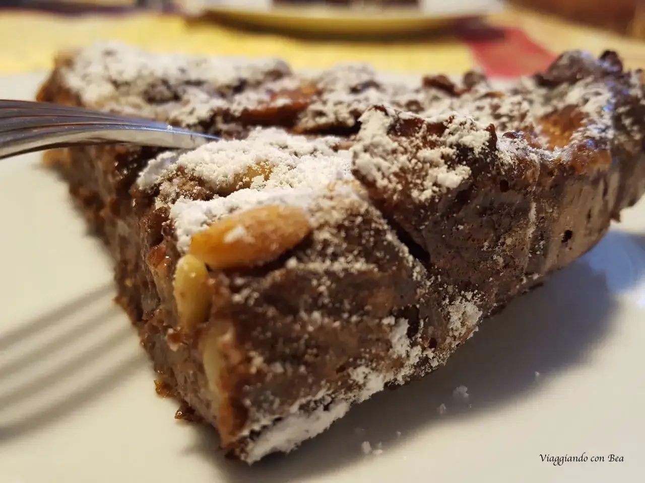 Una fetta di torta di pane ricetta della nonna, cosparsa di zucchero a velo e pinoli, pronta per essere gustata con una forchetta.