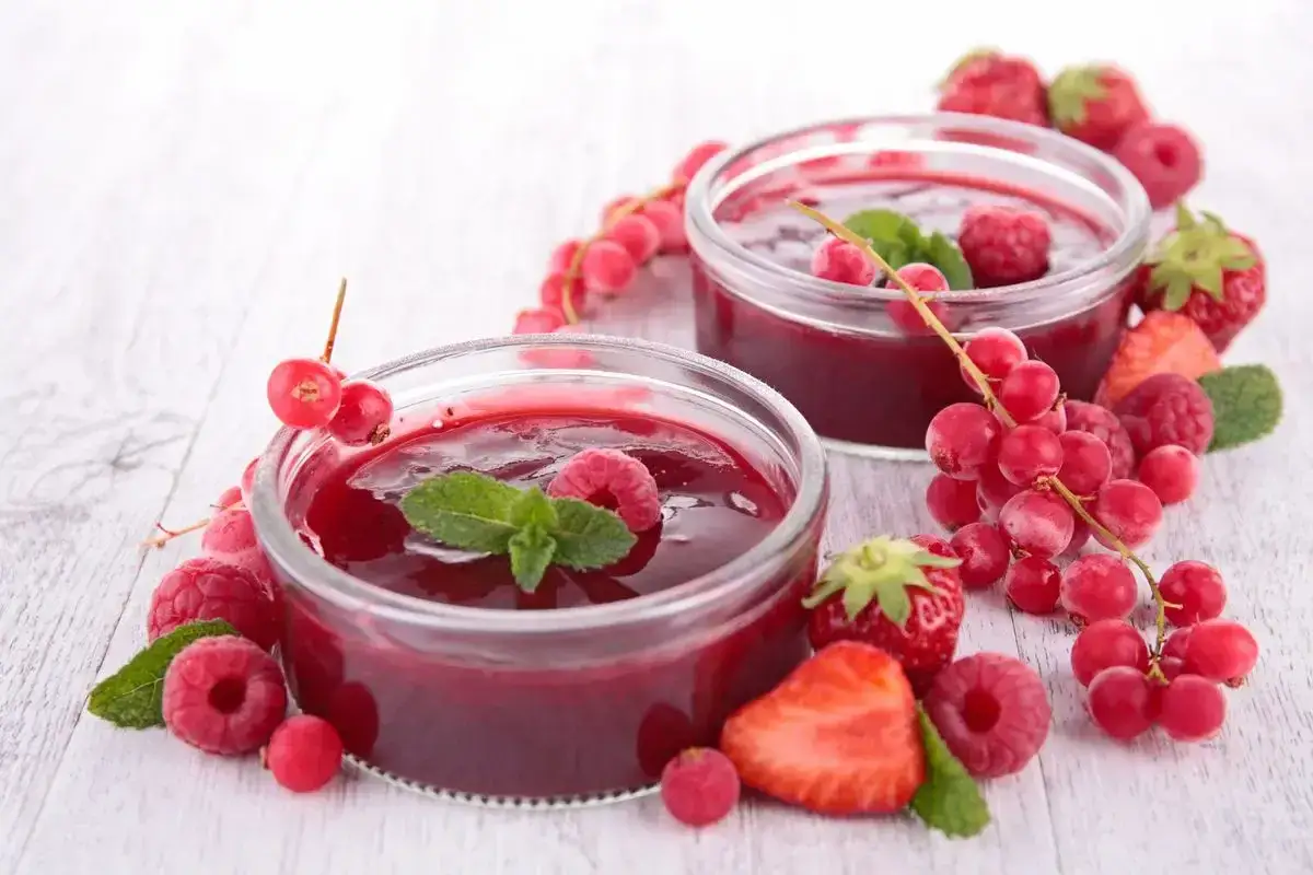 Due coppette di coulis di lamponi, guarnite con lamponi freschi, ribes e foglie di menta, circondate da fragole.