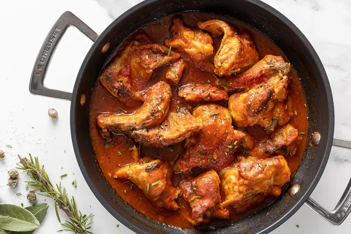 Un saporito coniglio in padella, cotto in un sugo rosso ricco, guarnito con erbe aromatiche.