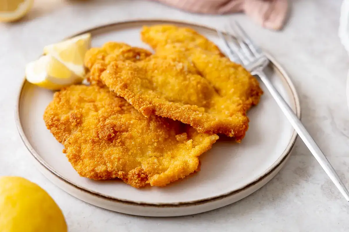 Piatto di cotolette di pollo dorate e croccanti, servite con spicchi di limone fresco e una forchetta.