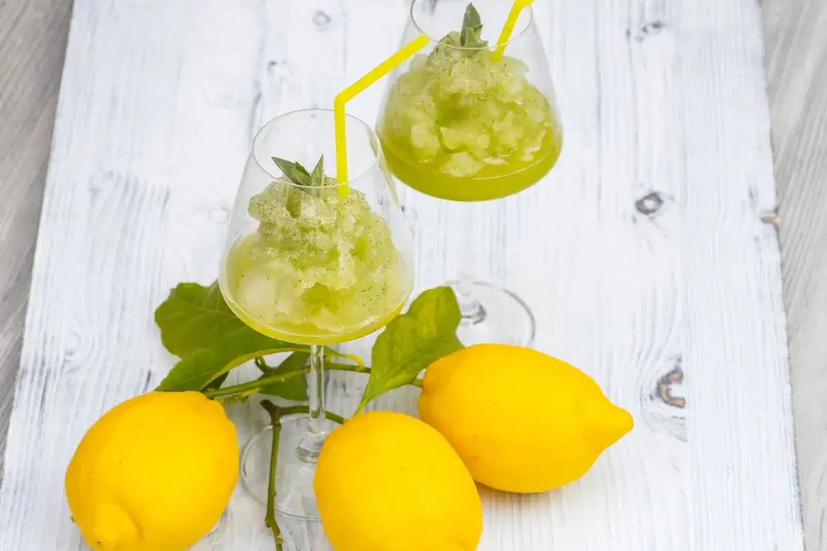 Due bicchieri di sorbetto al limone senza albume, guarniti con menta e cannucce gialle, accanto a limoni freschi su un tavolo di legno.