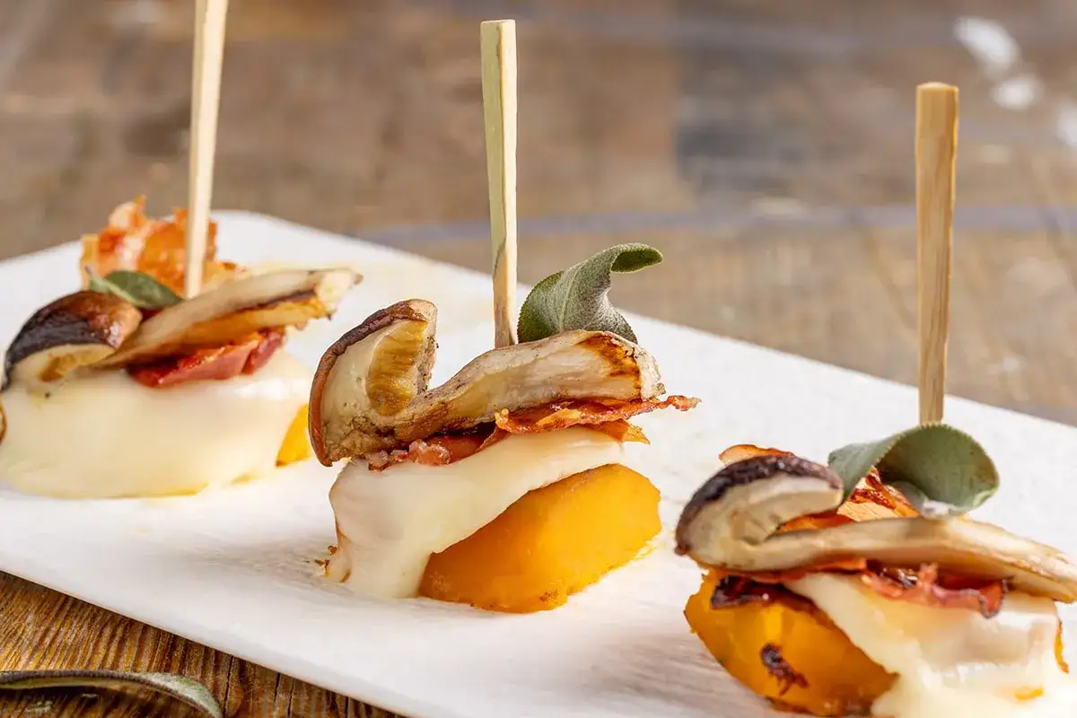 Antipasti sfiziosi per stupire: cubetti di zucca, formaggio filante, funghi e pancetta croccante, guarniti con salvia.