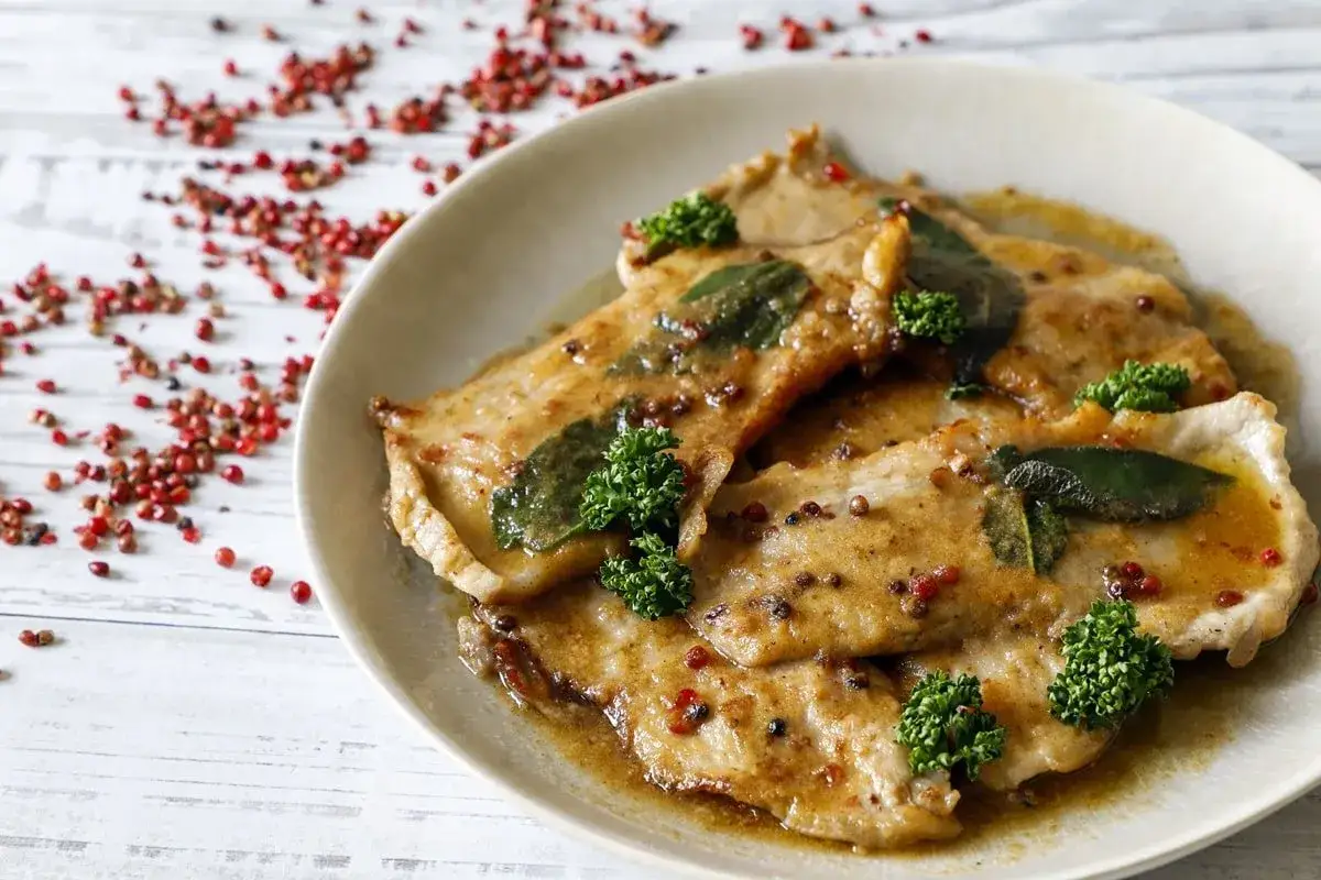 Scaloppine al vino bianco succulente, guarnite con salvia e prezzemolo, su un piatto con pepe rosa sparso.