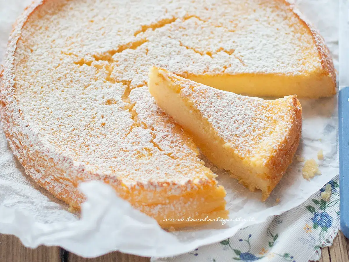 A slice of ricotta cake dusted with powdered sugar sits next to the whole cake.