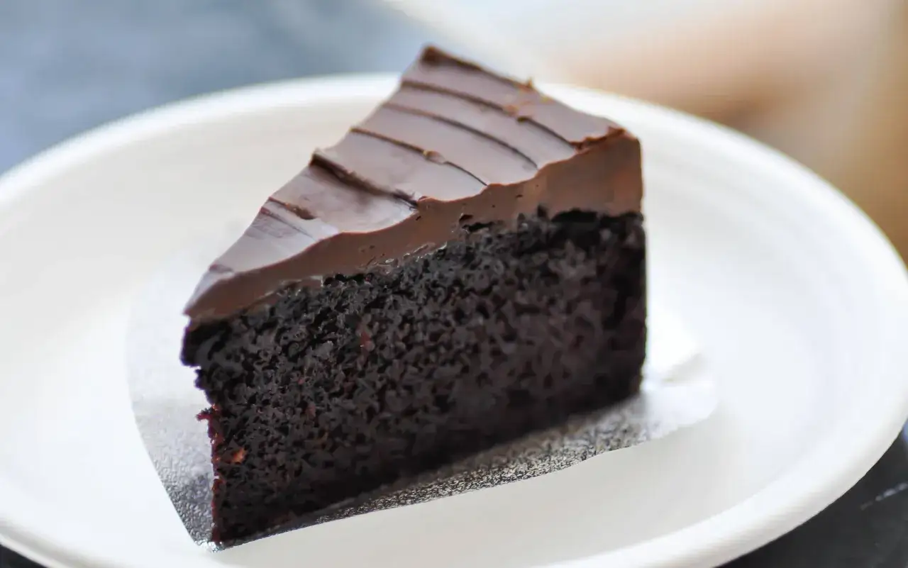 Una fetta di torta al cioccolato senza burro, con glassa lucida e un impasto soffice e scuro, servita su un piatto bianco.