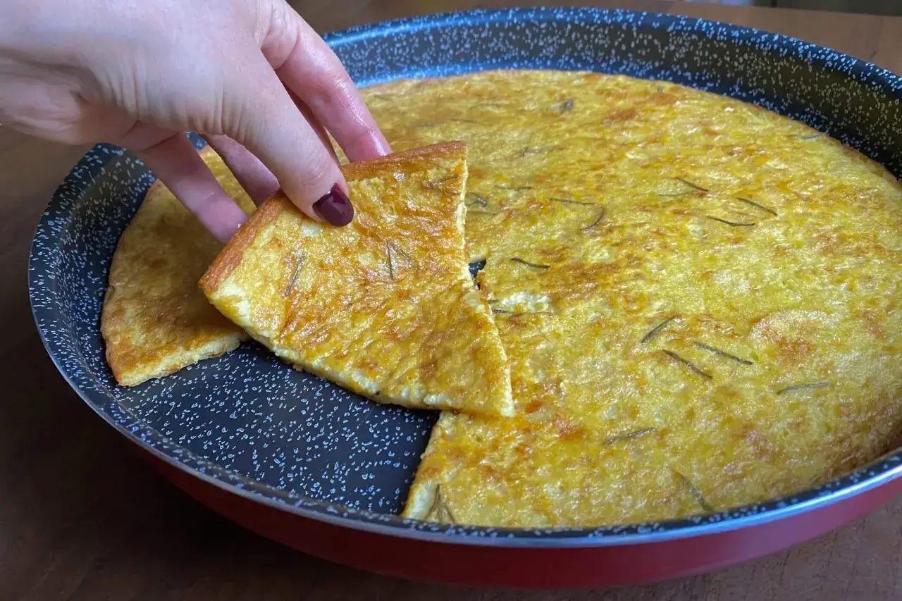 Una mano prende una fetta di dorata farinata di ceci, profumata al rosmarino, appena sfornata in una teglia.