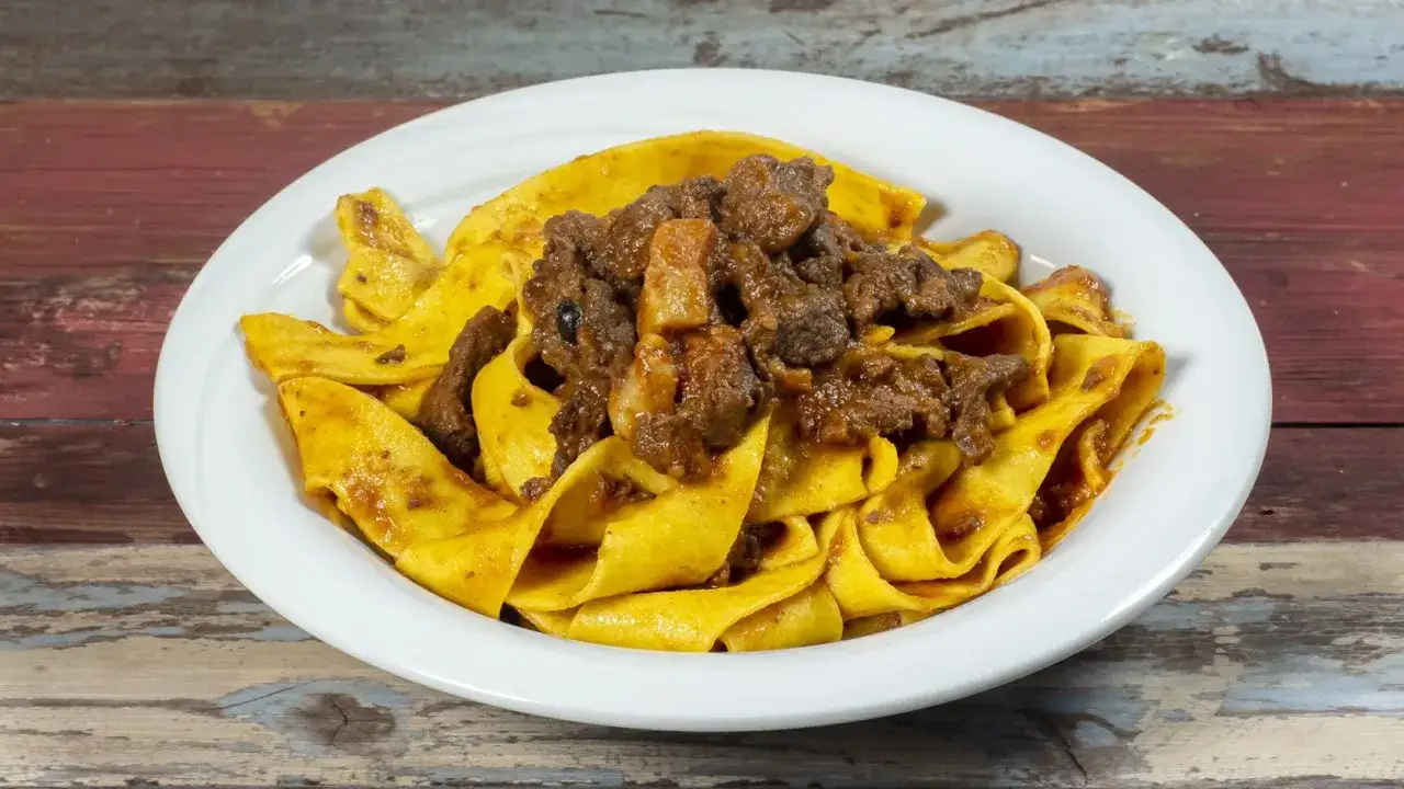 Pappardelle con rag&ugrave; di cinghiale, un piatto ricco e saporito, servito in un piatto bianco su un tavolo di legno.