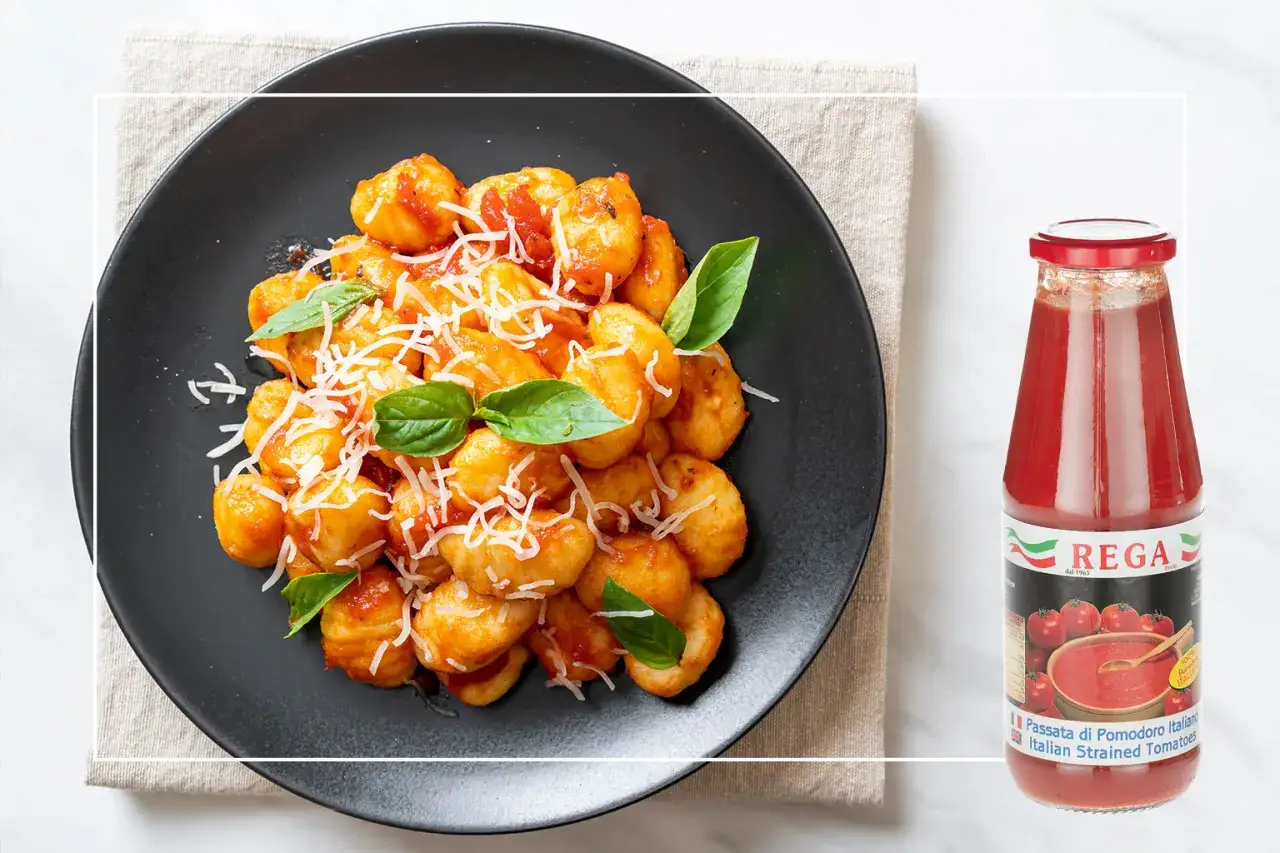 Gnocchi alla sorrentina fumanti su piatto nero, conditi con sugo di pomodoro e parmigiano, accanto a una bottiglia di passata Rega.