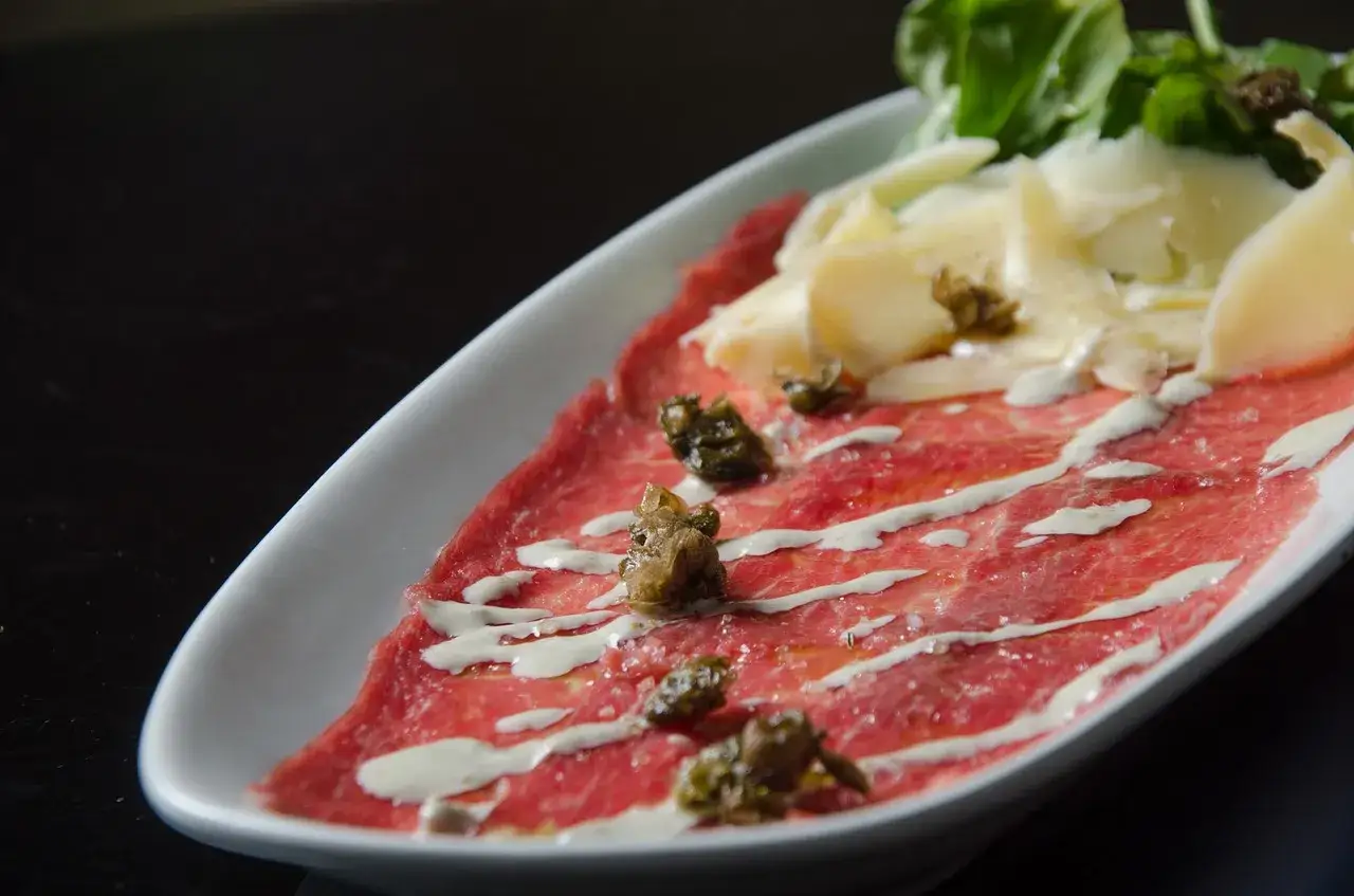 Delicato carpaccio di manzo con scaglie di parmigiano, capperi e un filo di salsa bianca su un piatto ovale bianco.