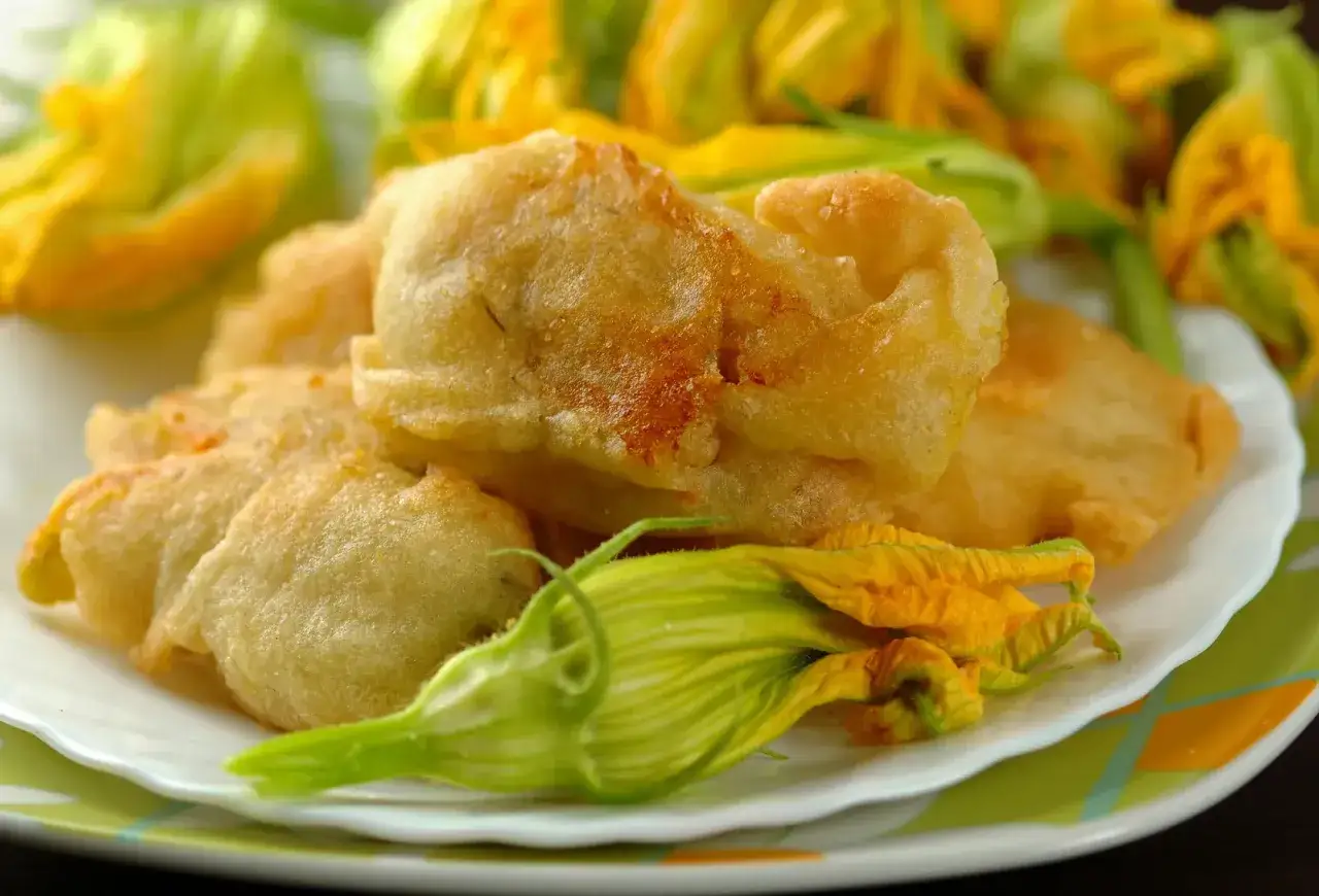 Fiori di zucca in padella, dorati e croccanti, serviti su un piatto bianco con fiori freschi sullo sfondo.