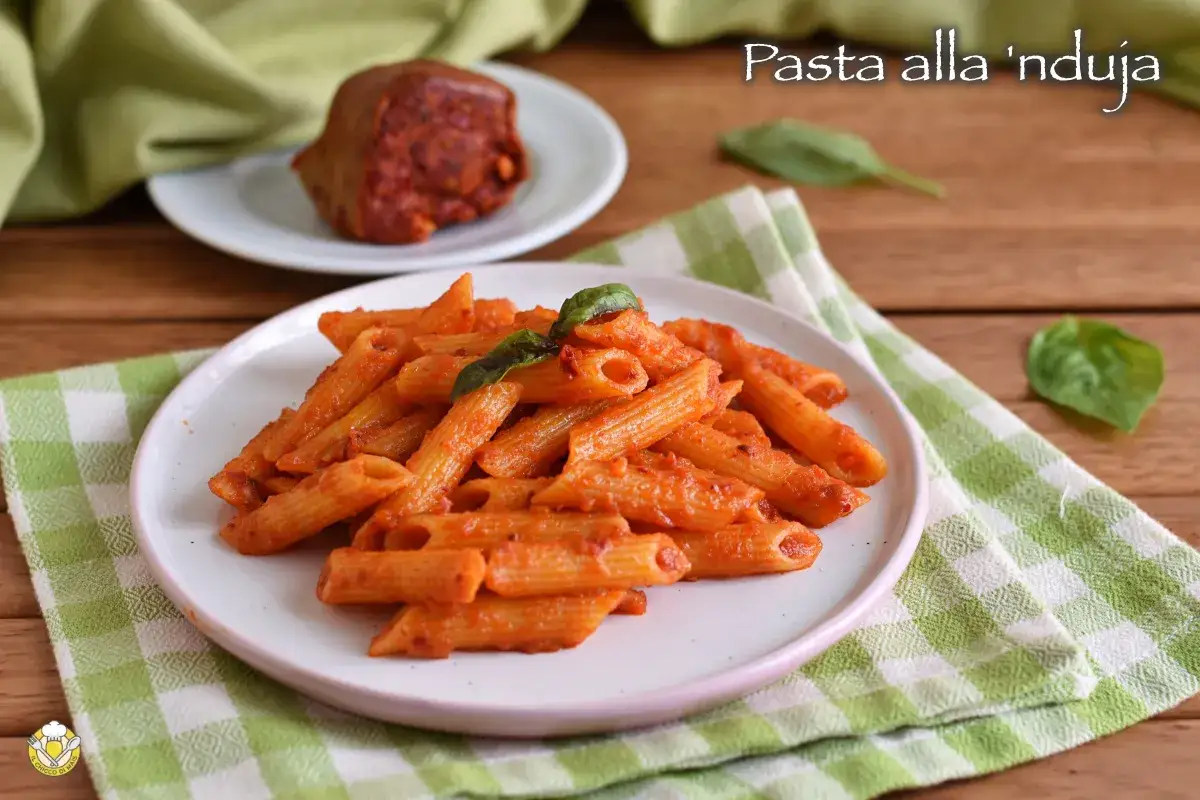 Piatto di penne al sugo rosso piccante, con un pezzo di 'nduja sullo sfondo.