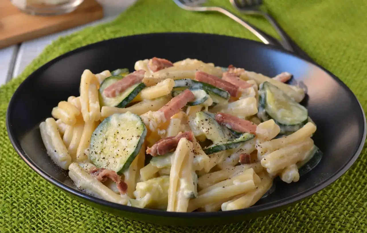 Piatto di pasta cremosa con zucchine a rondelle e cubetti di speck croccante, condita con pepe nero.