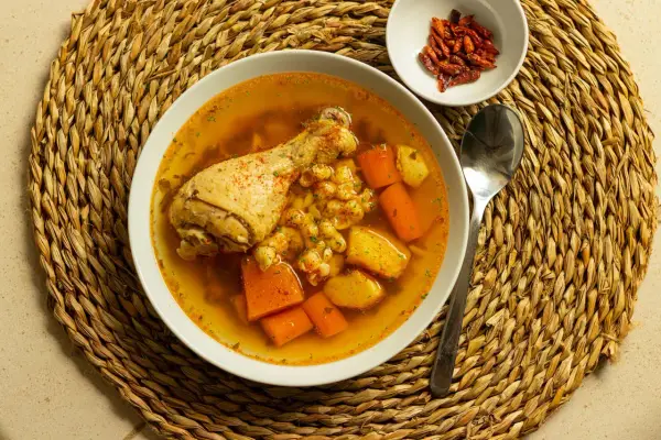 Un confortante brodo di pollo con pezzi di pollo, carote e patate, servito su una tovaglietta di paglia.